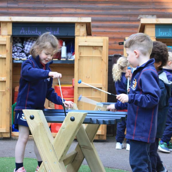 two school pupils playing with outdoor Xylophone in playground with storage units in the background