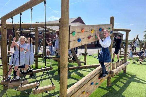 Children enjoying a new playground at Bury Grammar School featuring a wooden climbing wall and rope bridges on artificial turf, enhancing their physical activity and outdoor play.