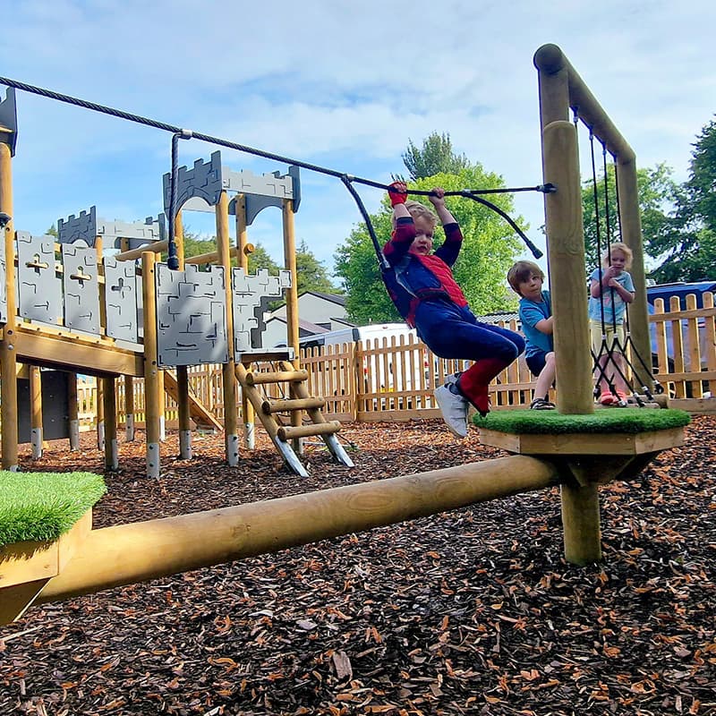 child in a spiderman costume swinging on a trim trail rope in play area with a wood chip surface and Pentagon commercial play tower in the background