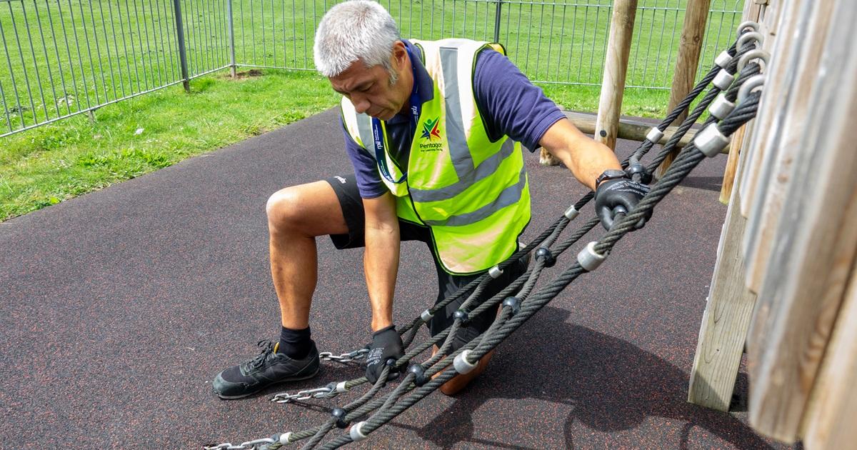 A man is holding on to a climbing rope net that is attached to a play tower and is testing the strength of it during a playground inspection