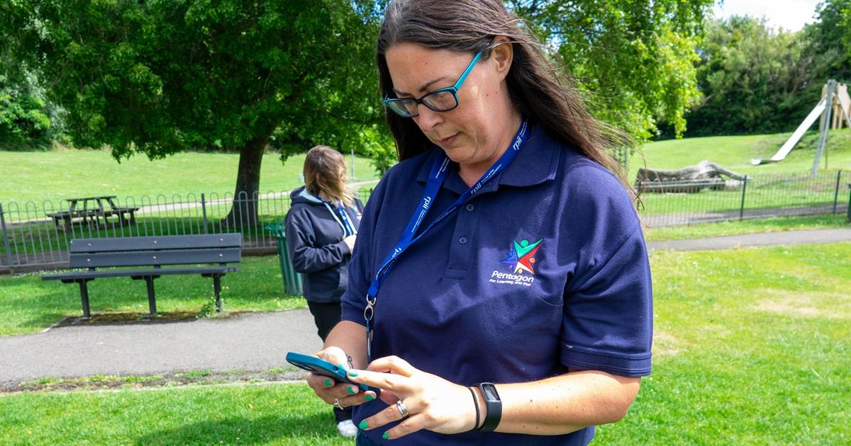 A playground inspector is holding her phone and is dialling a number to call a client, whilst she is stood on the playground