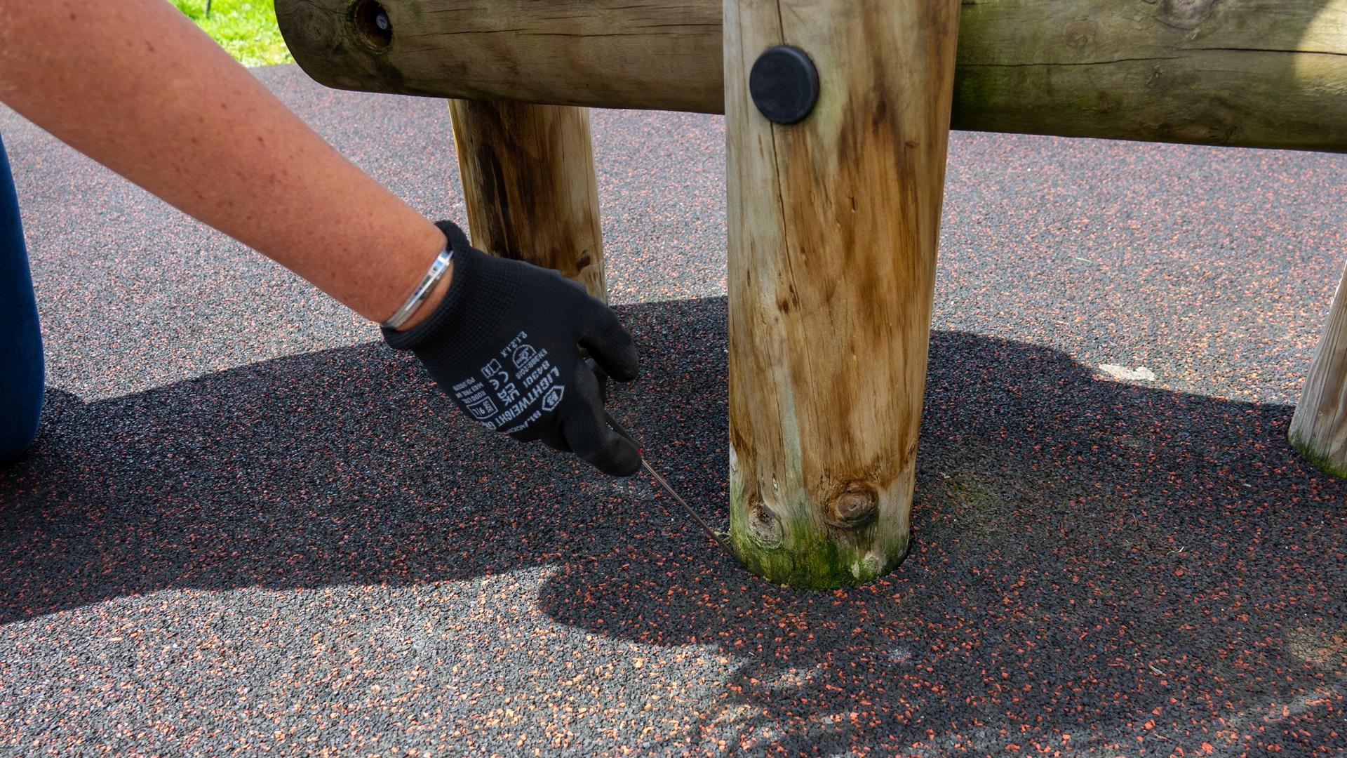 A playground inspector is holding a screwdriver as she prys away the wetpour that surrounds a wooden beam, showcasing the mold and rot that is affecting the piece of play equipment