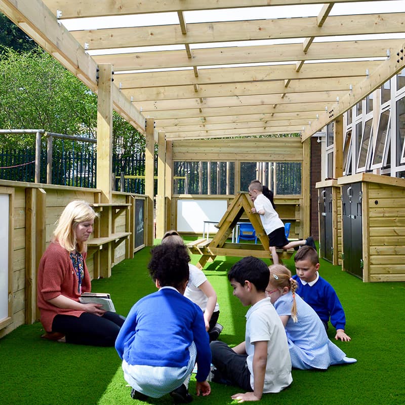 teacher working with a small group of students sat down on the astroturf in school canopy against main school building