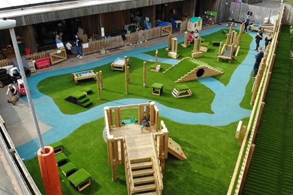 Mill Water School’s new playground featuring various wooden play structures on artificial turf, including ramps, tunnels, and a climbing area, designed for inclusive, active play.
