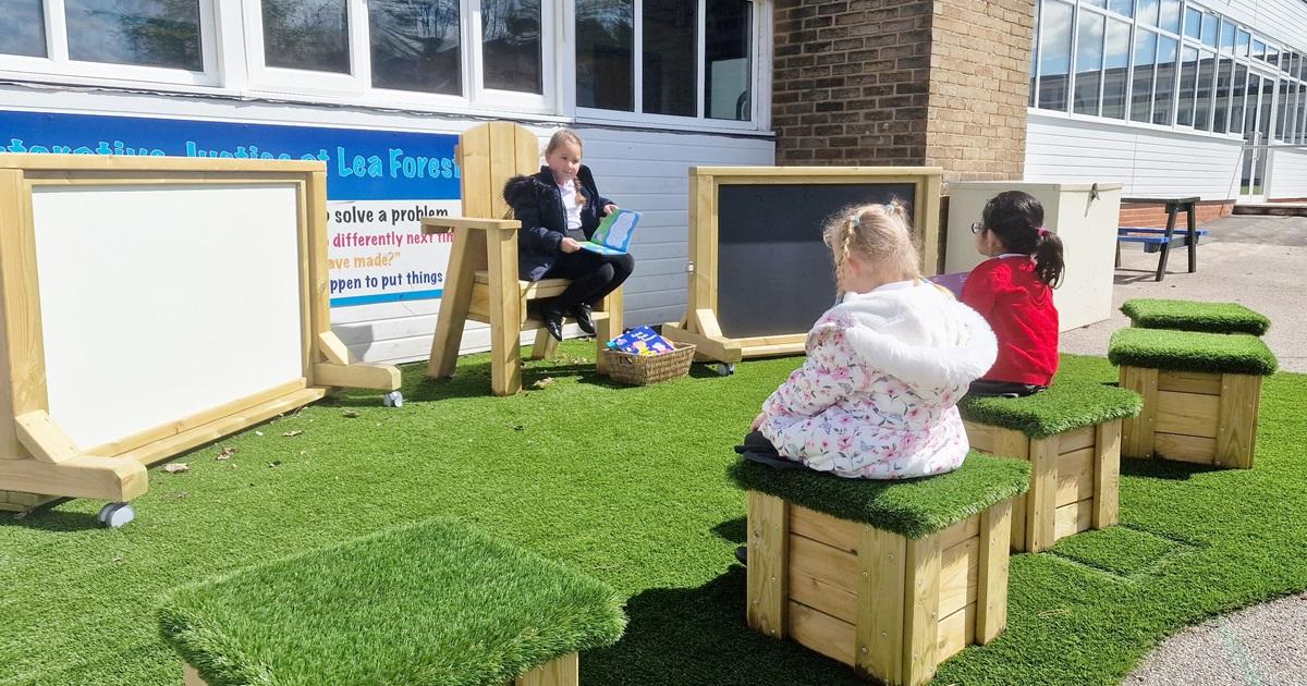 primary school child sitting on storytelling chair reading to friends sat on artificial grass-topped seats