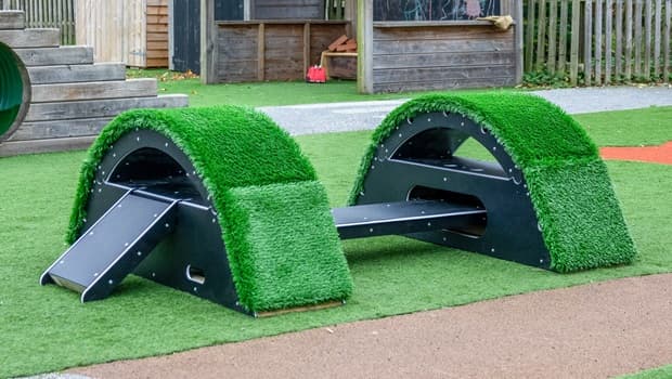 Two small grass-covered bridges with a black plank connecting them, outside in a school playground area