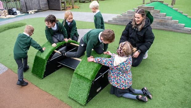 Two small grass-covered bridges with a black plank connecting them, with 6 school children playing on top and a female teacher watching them