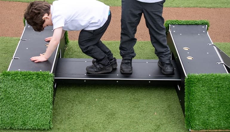 Two small grass-covered bridges with a black plank connecting them, with two school boys balancing on the black plank