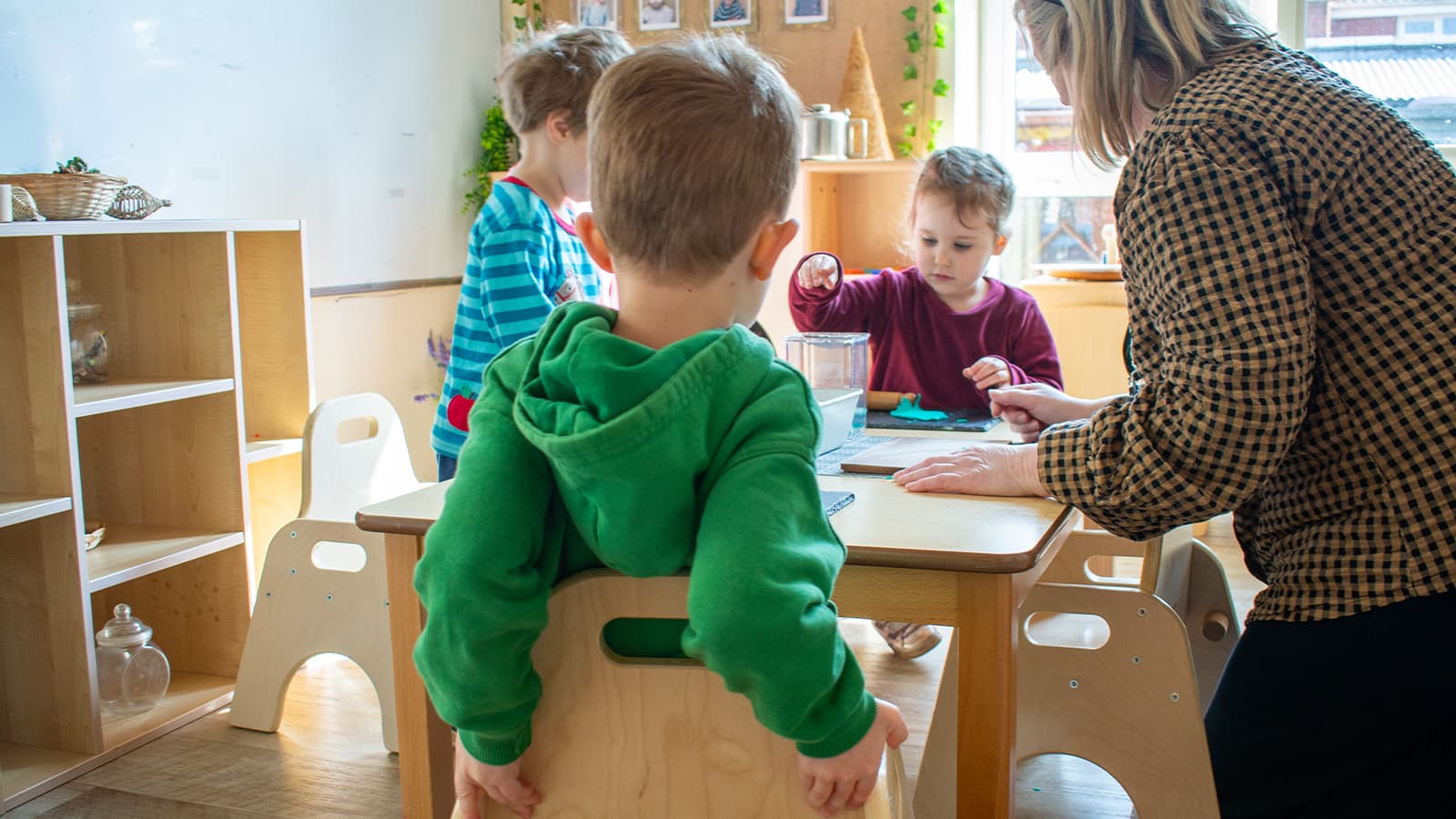A female teacher and three toddlers playing round a wooden table inside a nursery