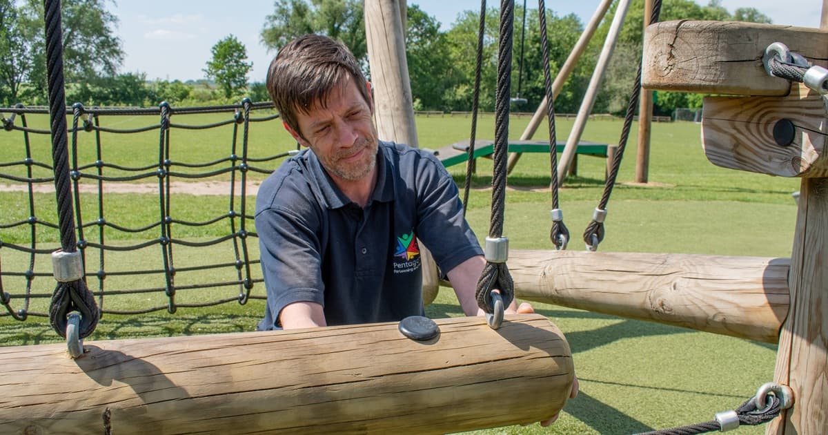 A man is holding on to a climbing rope net that is attached to a play tower and is testing the strength of it during a playground inspection
